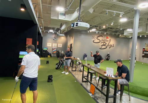 Inside a swing bays facility where golfers are hitting golf balls at simulator bays, and people are standing behind watching and talking.